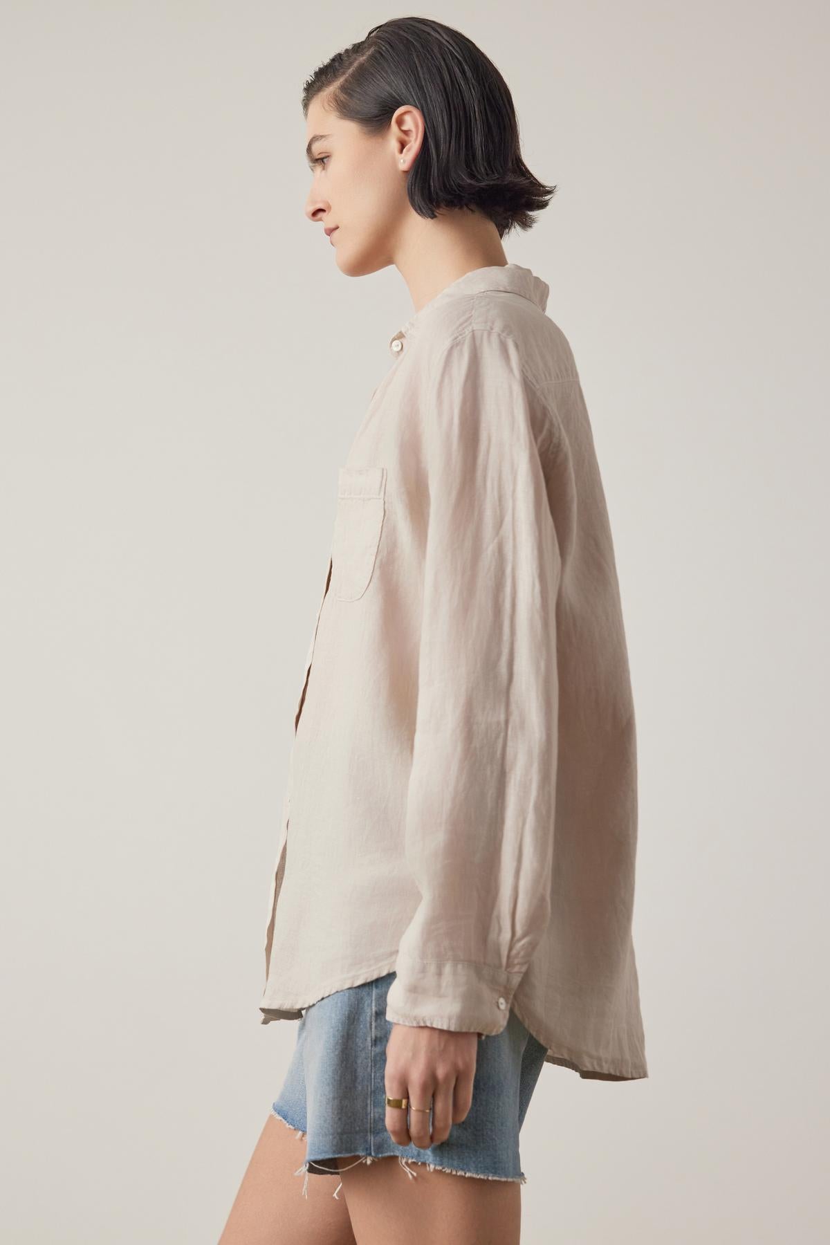   A person with short hair is wearing a loose, long-sleeve beige Velvet by Jenny Graham MULHOLLAND LINEN SHIRT and denim shorts, standing sideways against a plain background. The relaxed silhouette and scooped hemline of the shirt add to the casual vibe. 