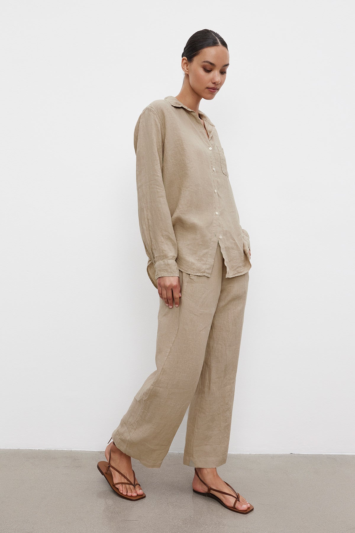   A person stands with a relaxed silhouette, wearing the MULHOLLAND LINEN SHIRT by Velvet by Jenny Graham, a matching beige linen button-up shirt with a scooped hemline and wide-leg pants, paired with brown sandals. The background is a plain white wall and grey floor. 