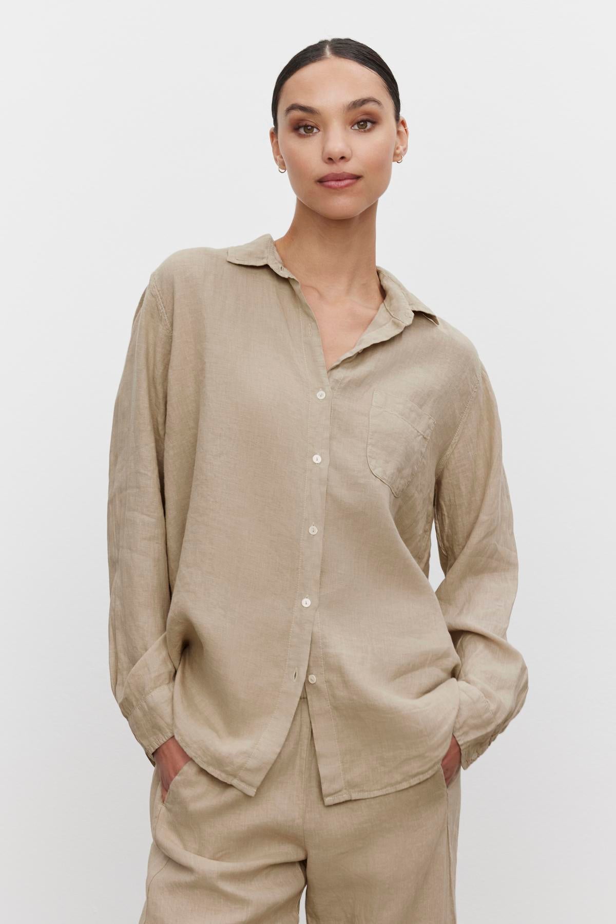   A person wearing the MULHOLLAND LINEN SHIRT in light beige by Velvet by Jenny Graham, paired with matching pants, stands against a plain white background. Their hands are in their pockets, and they have a slight smile, exuding a relaxed silhouette. 