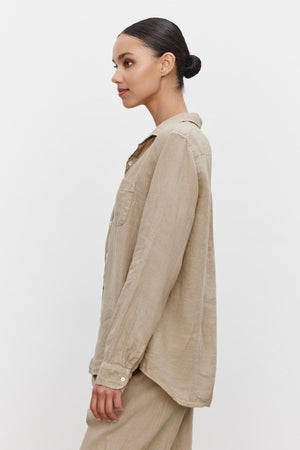 A person with dark hair tied in a bun is standing sideways, wearing a light beige linen button-up shirt and matching pants, featuring a scooped hemline against a plain white background. The shirt is the MULHOLLAND LINEN SHIRT by Velvet by Jenny Graham.