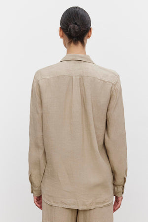 A person with hair in a bun is seen from the back, wearing a beige, long-sleeved MULHOLLAND LINEN SHIRT by Velvet by Jenny Graham with a relaxed silhouette, standing against a plain white background.