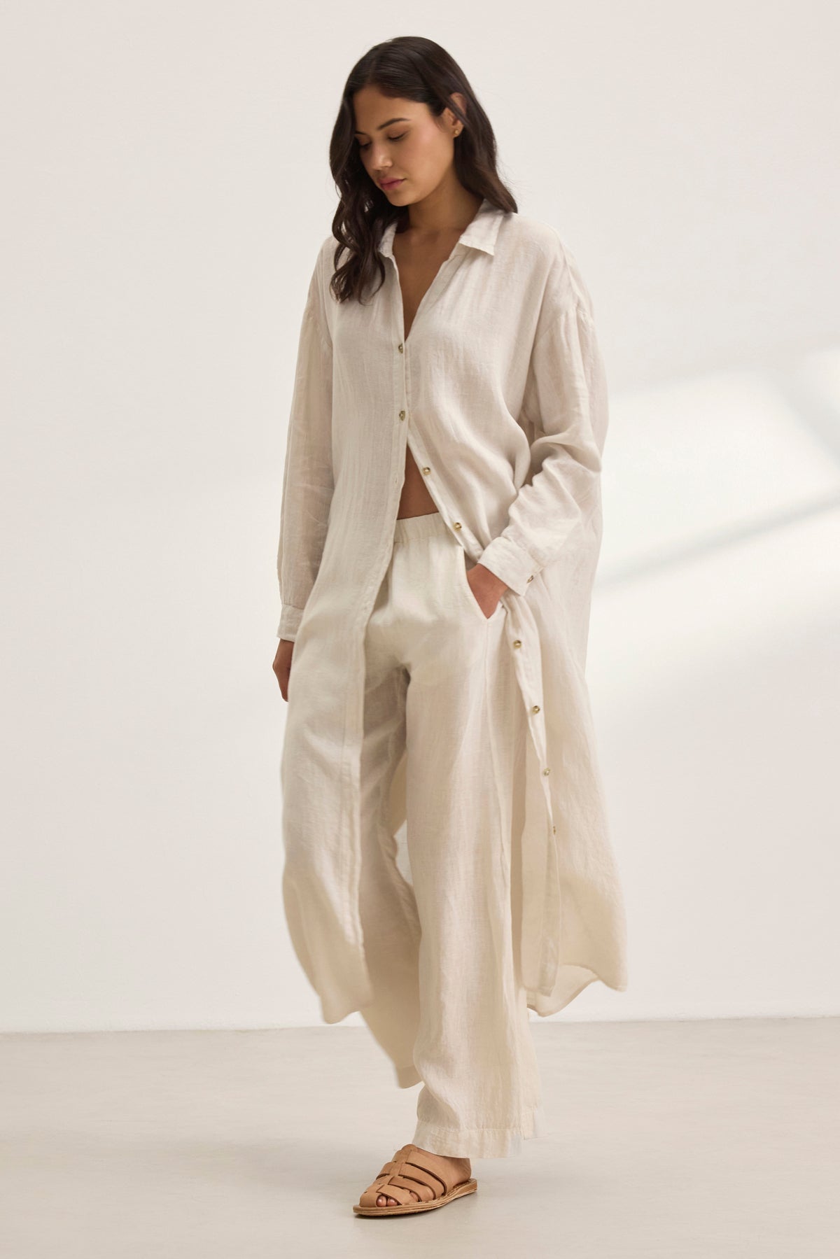   A woman wearing the Velvet by Graham & Spencer NICOLE LINEN DRESS and matching wide-leg pants stands in a neutral room with soft shadows, embodying breathable comfort. 