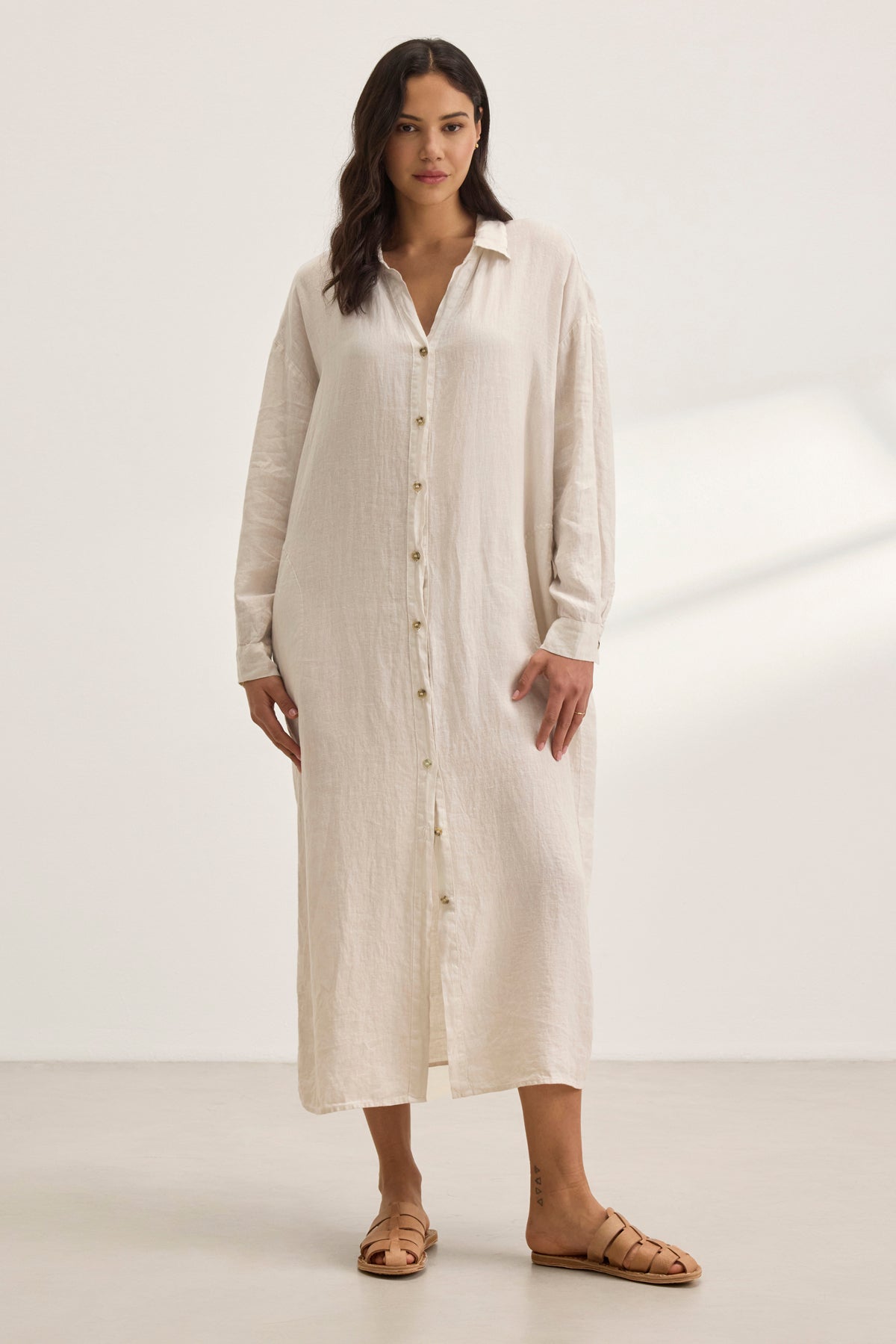 A woman elegantly wears the NICOLE LINEN DRESS by Velvet by Graham & Spencer, a long, buttoned shirt dress in light beige woven linen. Its side seam patch pockets enhance both style and utility amidst a neutral setting.-38819751329985