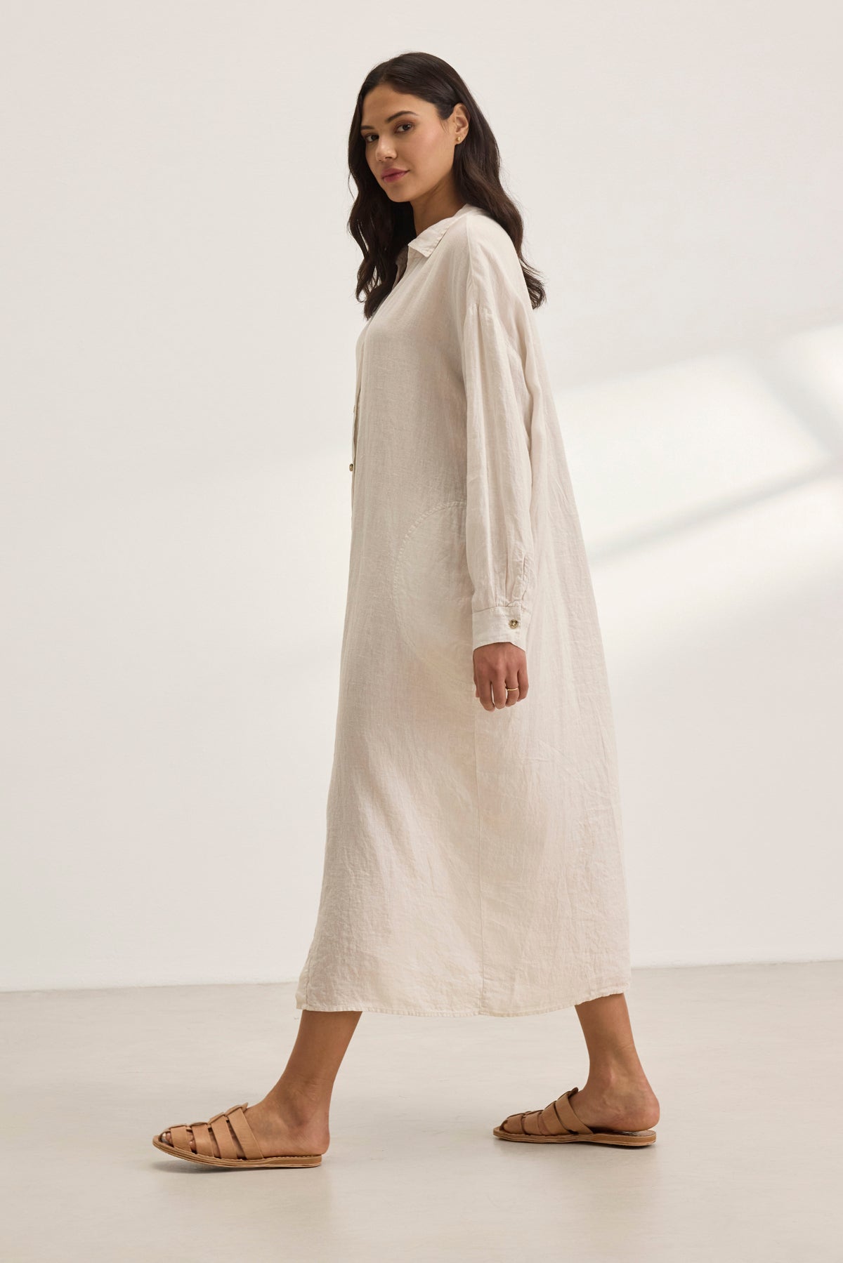   A woman in a NICOLE LINEN DRESS by Velvet by Graham & Spencer strides forward against a plain background, embodying breathable comfort with every step, paired with sandals. 