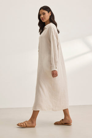 A woman in a NICOLE LINEN DRESS by Velvet by Graham & Spencer strides forward against a plain background, embodying breathable comfort with every step, paired with sandals.