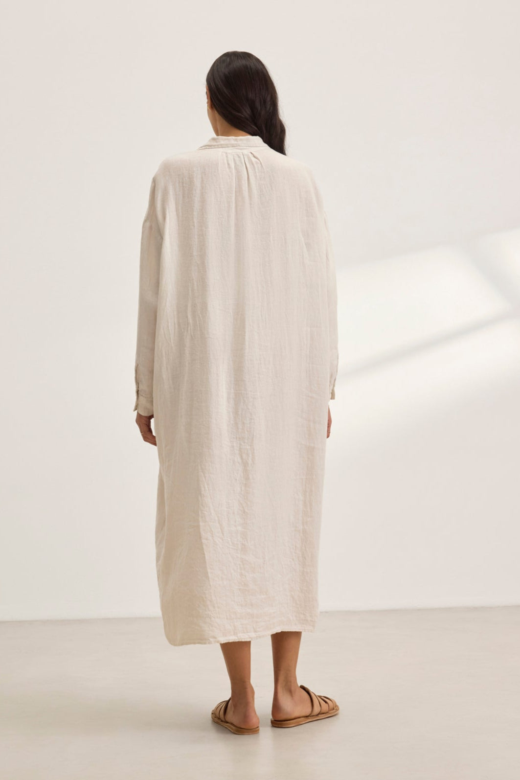   A person with long hair, barefoot, is wearing the cream-colored, versatile NICOLE LINEN DRESS by Velvet by Graham & Spencer while facing a plain white wall. 