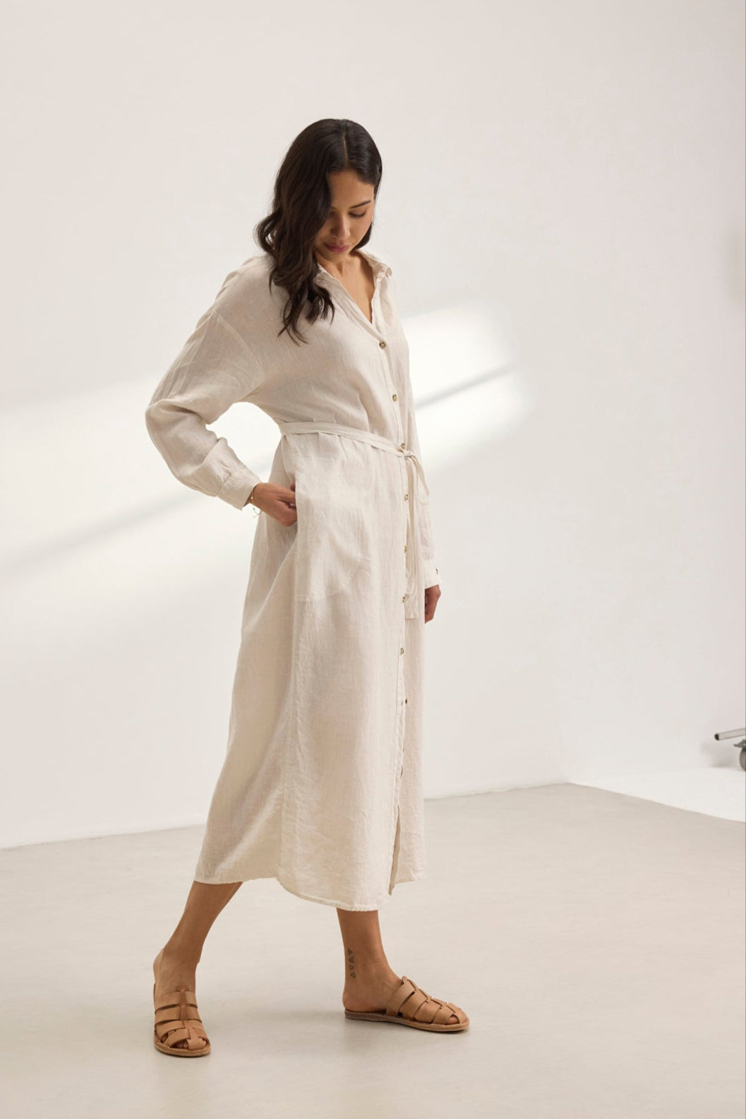   In a minimalist room, a person in the NICOLE LINEN DRESS by Velvet by Graham & Spencer pairs the versatile beige linen shirt dress with long sleeves and brown sandals, exuding breathable comfort while gazing down. 