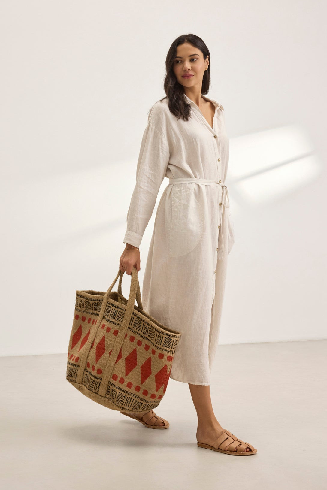   A woman embodies breathable comfort in a versatile NICOLE LINEN DRESS from Velvet by Graham & Spencer and sandals, holding a large decorative tote bag. 