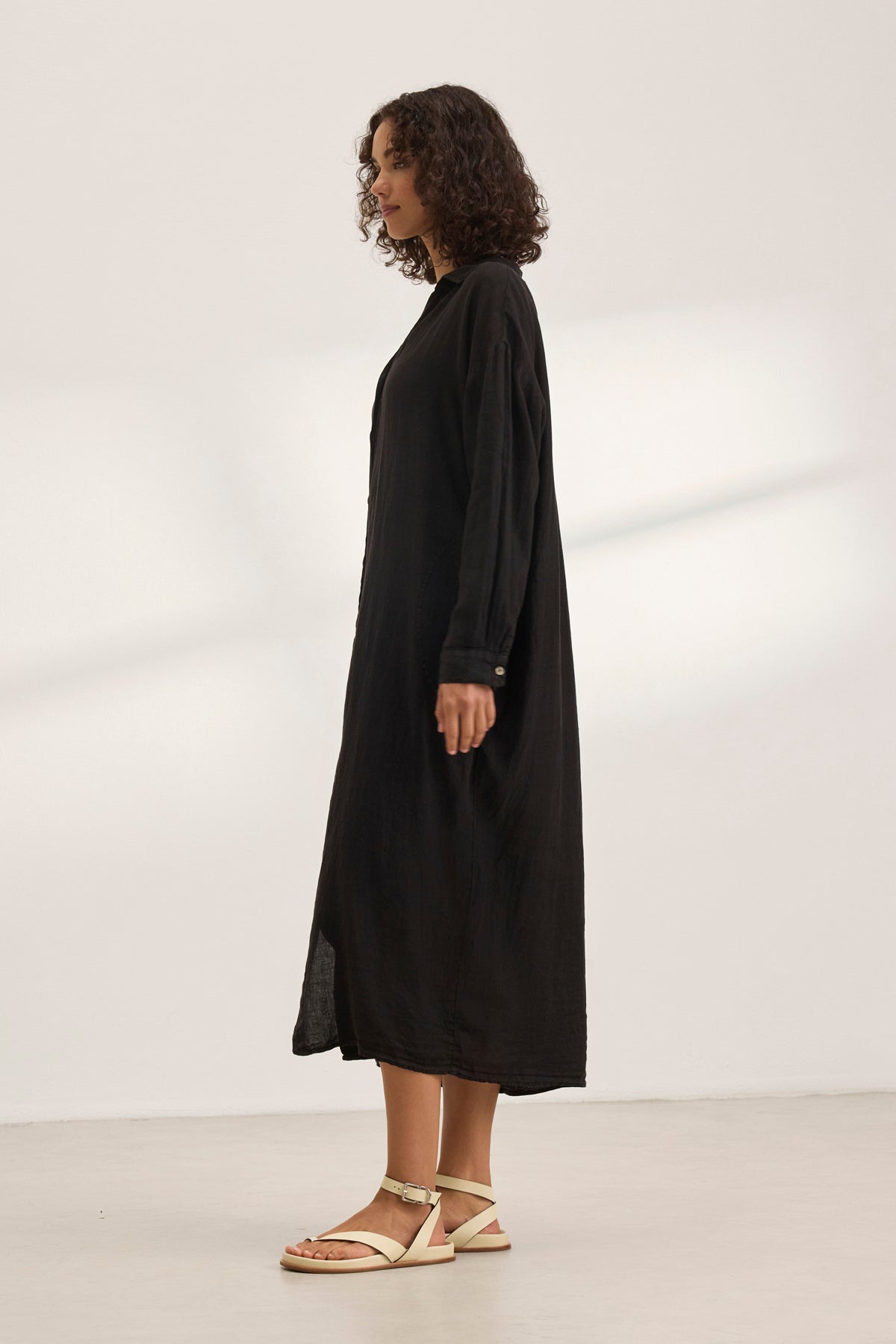   A woman in profile wears the NICOLE LINEN DRESS by Velvet by Graham & Spencer—a long black button-up shirt dress—with white sandals against a plain light background. The lightweight woven linen ensures effortless style and breathable comfort. 