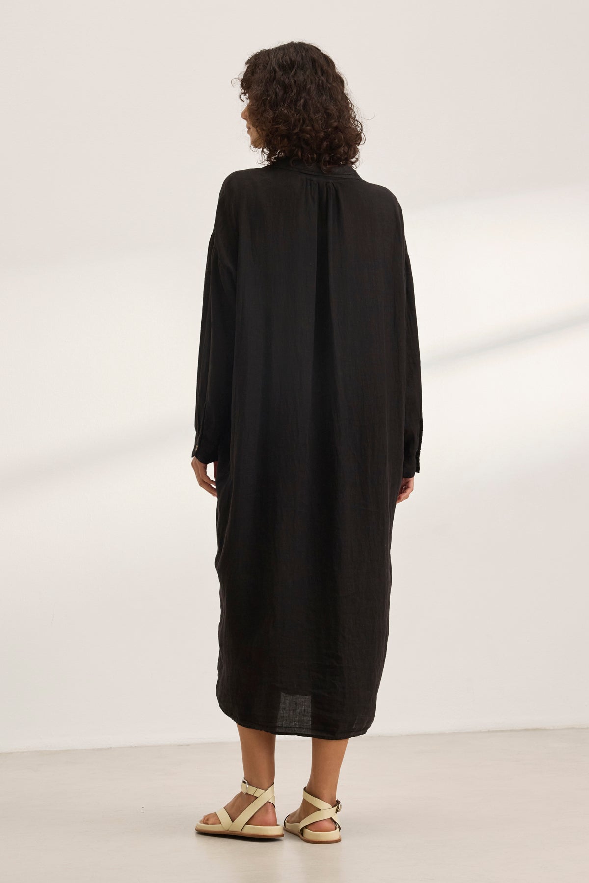   A person with curly hair is wearing the NICOLE LINEN DRESS by Velvet by Graham & Spencer and beige sandals, standing with their back to the camera in a minimalist white room. 