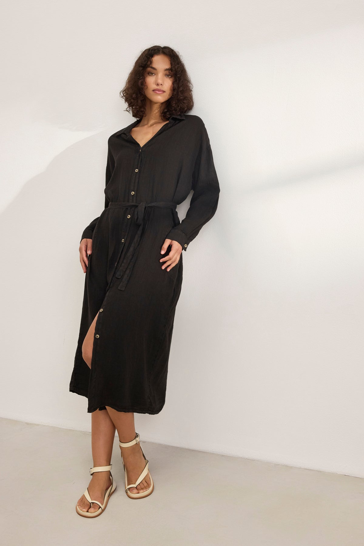   Wearing the versatile NICOLE LINEN DRESS in long black with buttons by Velvet by Graham & Spencer, paired with white sandals, a person stands against a plain light background. 