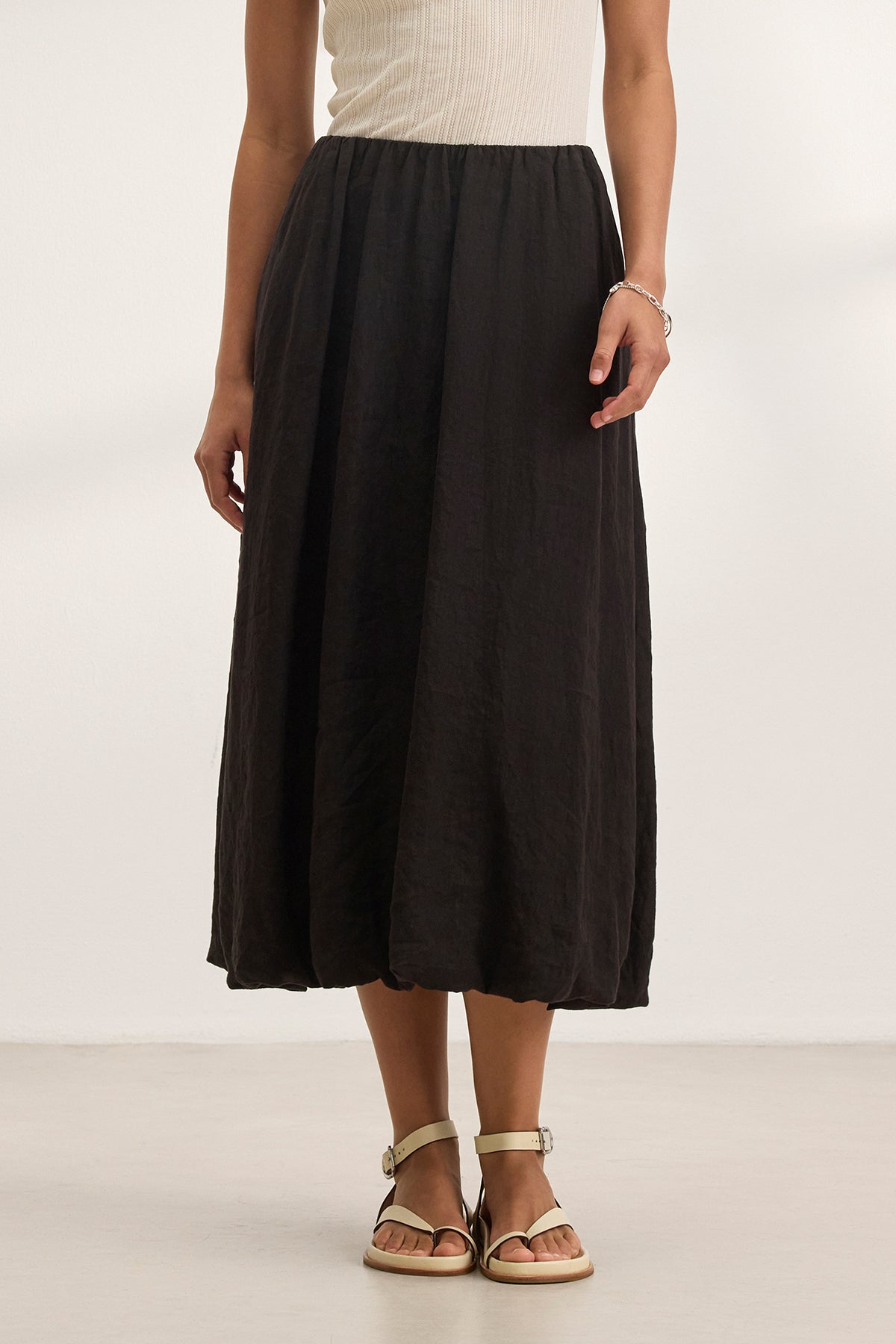   A person in a white sleeveless top, Velvet by Graham & Spencer's RACHELLE LINEN SKIRT in black bubble hem style, and tan sandals stands on a light-colored floor. 