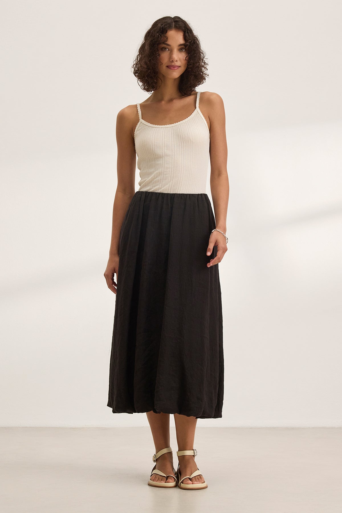   A woman wearing a white sleeveless top and the RACHELLE LINEN SKIRT by Velvet by Graham & Spencer, featuring a long black bubble hem design, stands against a plain background. 