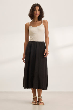 A woman wearing a white sleeveless top and the RACHELLE LINEN SKIRT by Velvet by Graham & Spencer, featuring a long black bubble hem design, stands against a plain background.