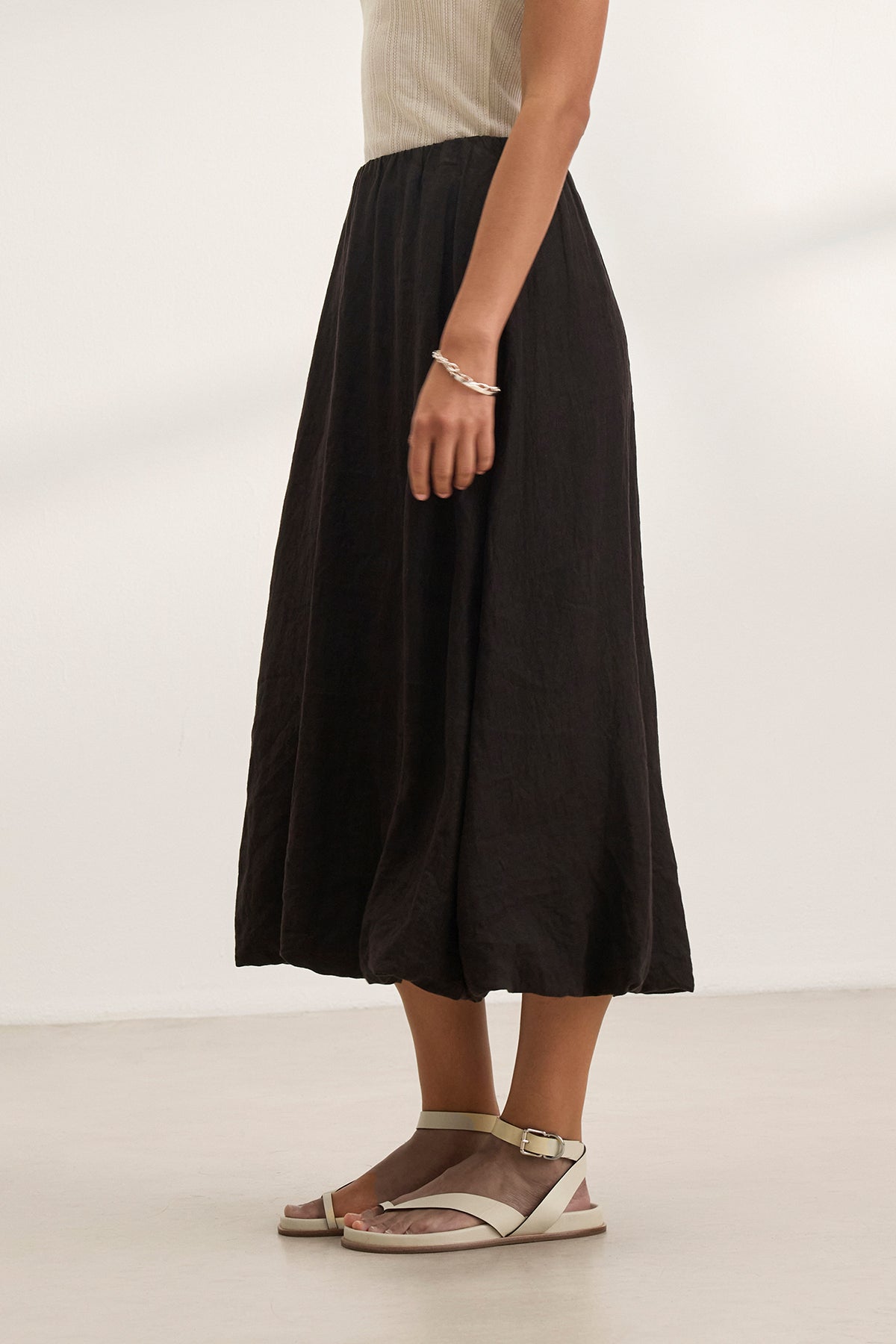   Sporting a sleeveless top, Velvet by Graham & Spencer's RACHELLE LINEN SKIRT in a long black bubble hem, and white sandals, the person stands against a plain background. 