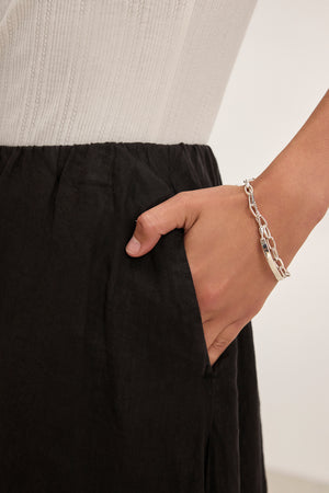 A person wearing a white top and Velvet by Graham & Spencer's black RACHELLE LINEN SKIRT with an elastic waist, hand in the pocket, showcasing a silver chain bracelet.