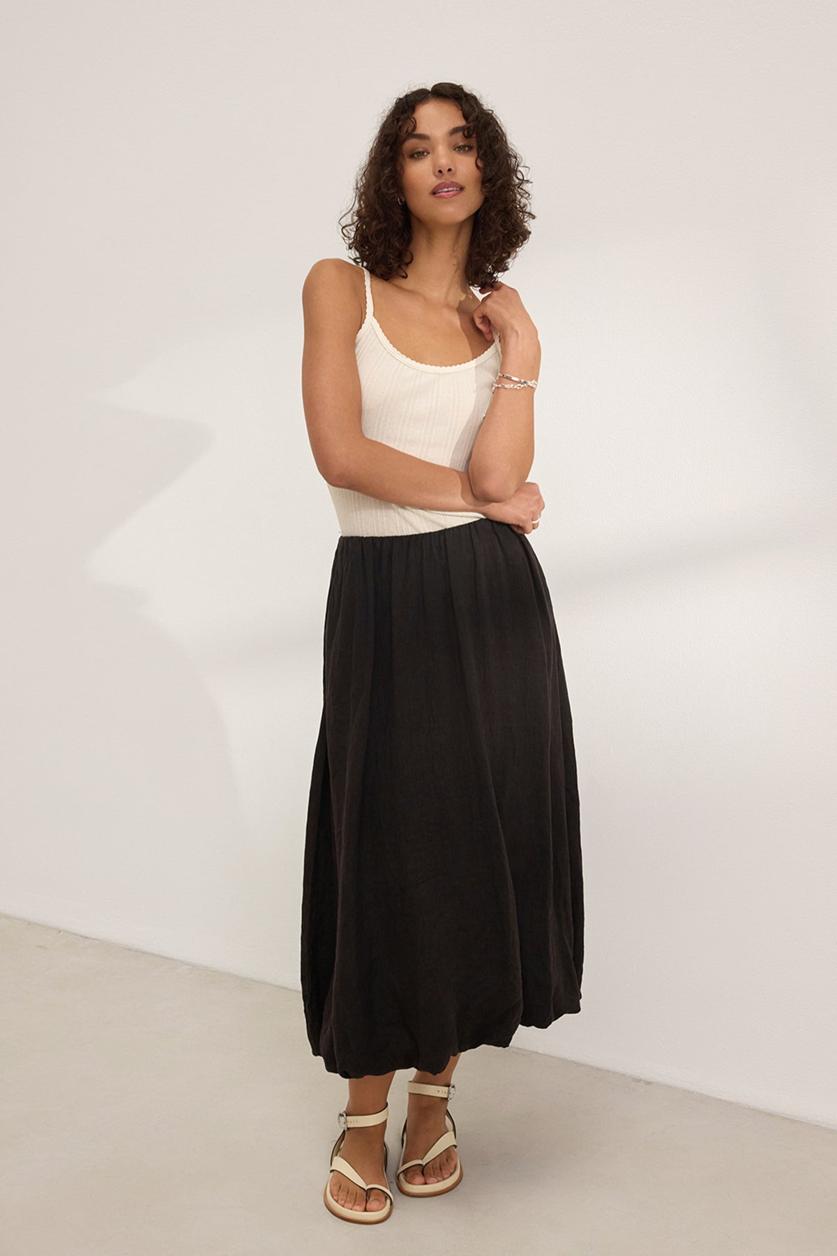   A woman with curly hair in a white tank top and the RACHELLE LINEN SKIRT by Velvet by Graham & Spencer stands against a plain white wall, her sandals completing the look. 