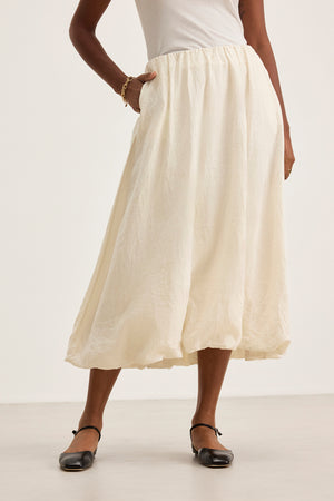 Someone in a white RACHELLE LINEN SKIRT by Velvet by Graham & Spencer, featuring a bubble hem, pairs it with a white top and black shoes, casually resting one hand in the skirt pocket.