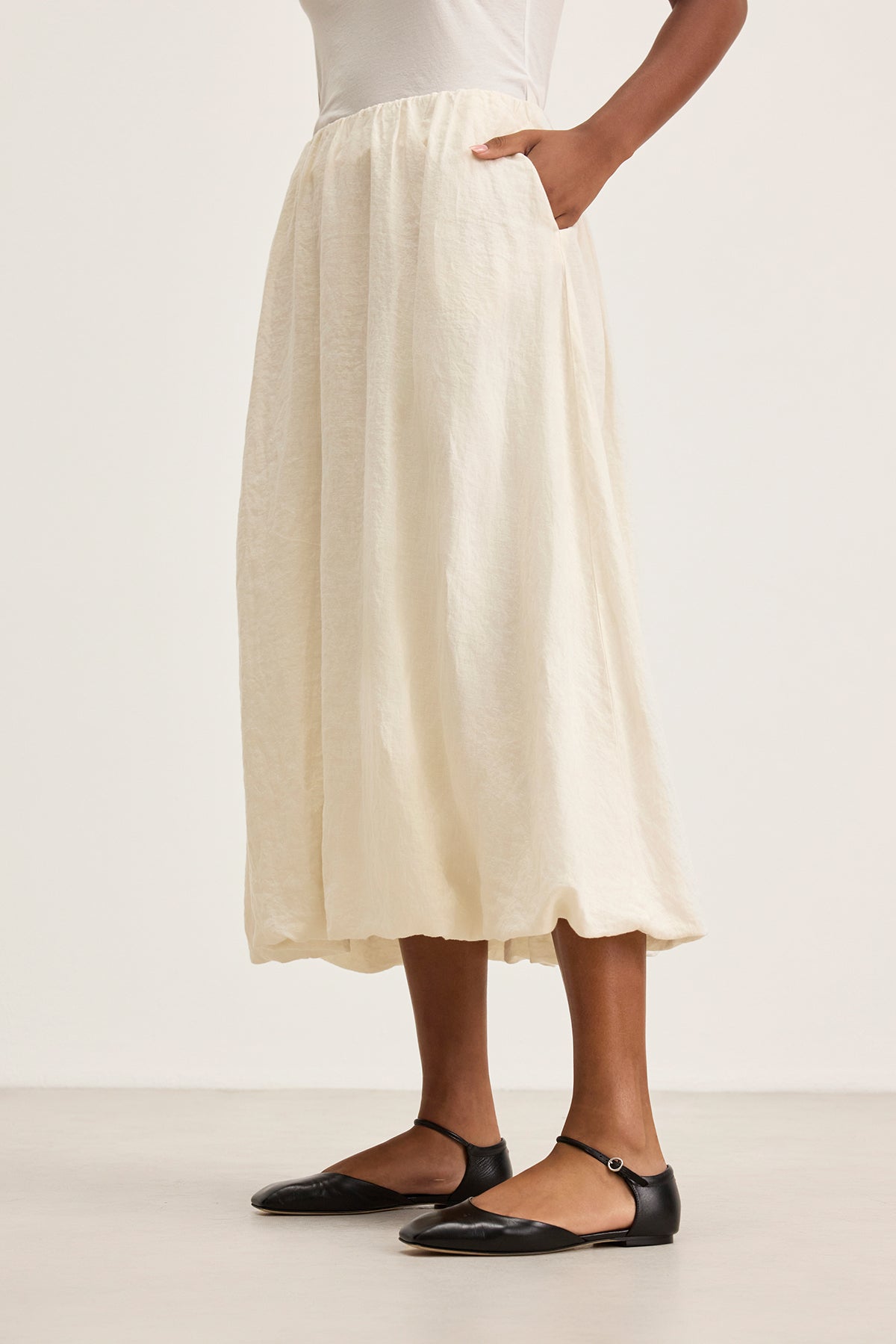   Someone is styled in the RACHELLE LINEN SKIRT by Velvet by Graham & Spencer, featuring a white, flowy design with an elastic waist and pockets, paired with black ballet flats against a neutral background. 