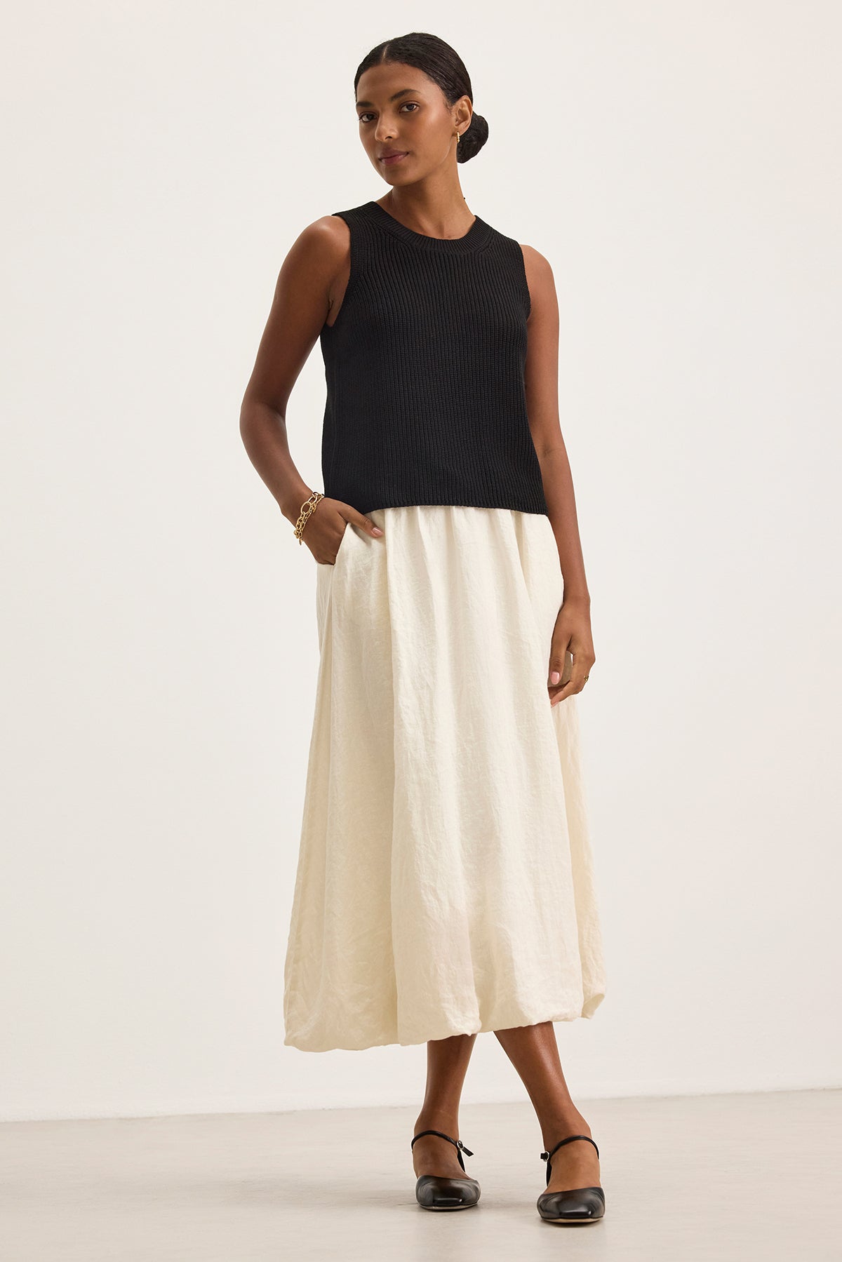 Against a plain background, a person wears a black sleeveless top and the RACHELLE LINEN SKIRT by Velvet by Graham & Spencer, featuring its long white design with a subtle bubble hem, while standing with one hand in the pocket.-38663160660161