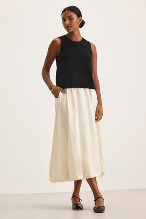 Against a plain background, a person wears a black sleeveless top and the RACHELLE LINEN SKIRT by Velvet by Graham & Spencer, featuring its long white design with a subtle bubble hem, while standing with one hand in the pocket.