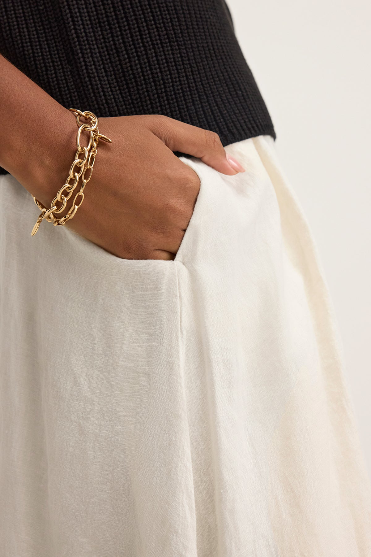   A person is wearing a black top, the RACHELLE LINEN SKIRT by Velvet by Graham & Spencer, featuring a white woven linen design with an elastic waist, and a gold chain bracelet, with their hand in the pocket. 