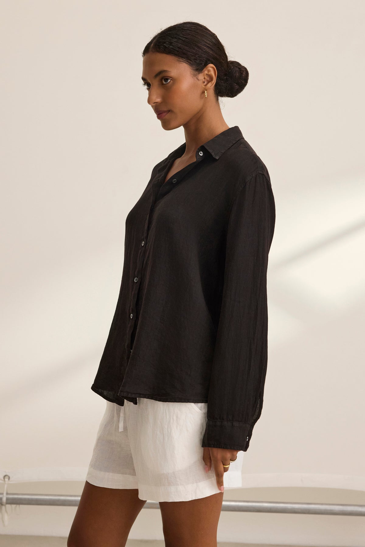 Against a neutral backdrop, someone stands in profile wearing the Velvet by Graham & Spencer SISI LINEN SHIRT and white shorts, exuding effortless style.-38819773907137