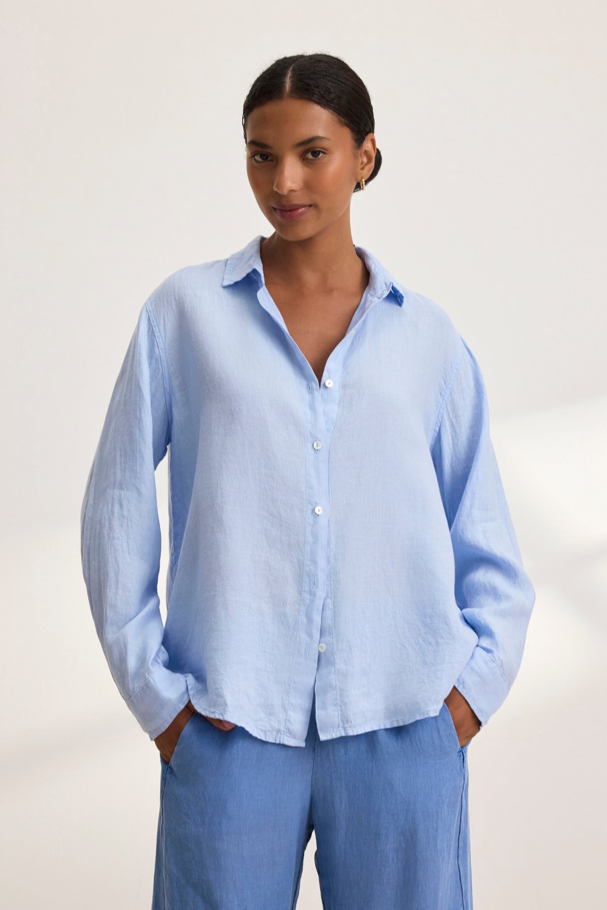   Person wearing a Velvet by Graham & Spencer SISI LINEN SHIRT in light blue, showcasing its breathable design, stands with hands in pockets against a plain background. 