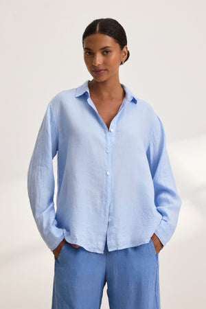 Person wearing a Velvet by Graham & Spencer SISI LINEN SHIRT in light blue, showcasing its breathable design, stands with hands in pockets against a plain background.