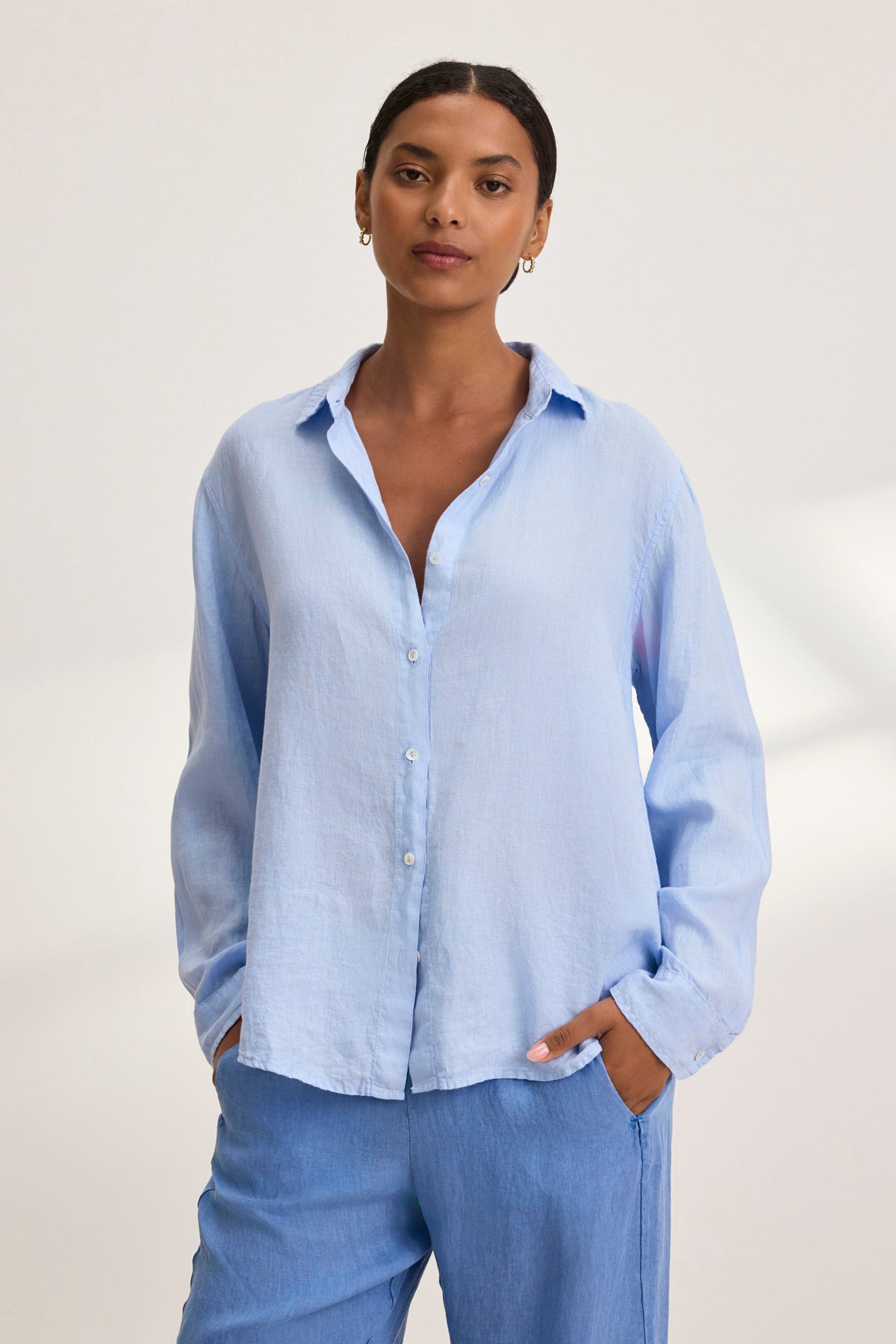   A person wearing a SISI LINEN SHIRT in light blue by Velvet by Graham & Spencer, paired with matching pants, stands with hands in pockets against a plain background. 