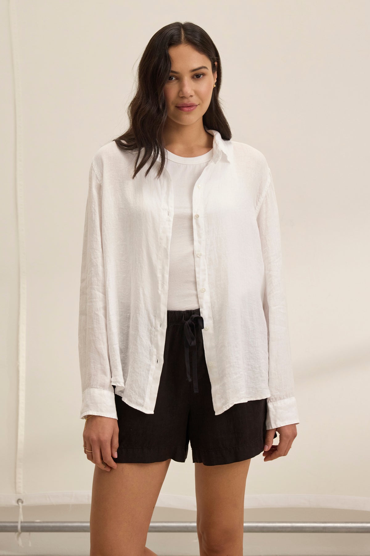   A woman stands against a plain background wearing the SISI LINEN SHIRT by Velvet by Graham & Spencer, a white button-up crafted from lightweight linen, over a white top and black shorts. 