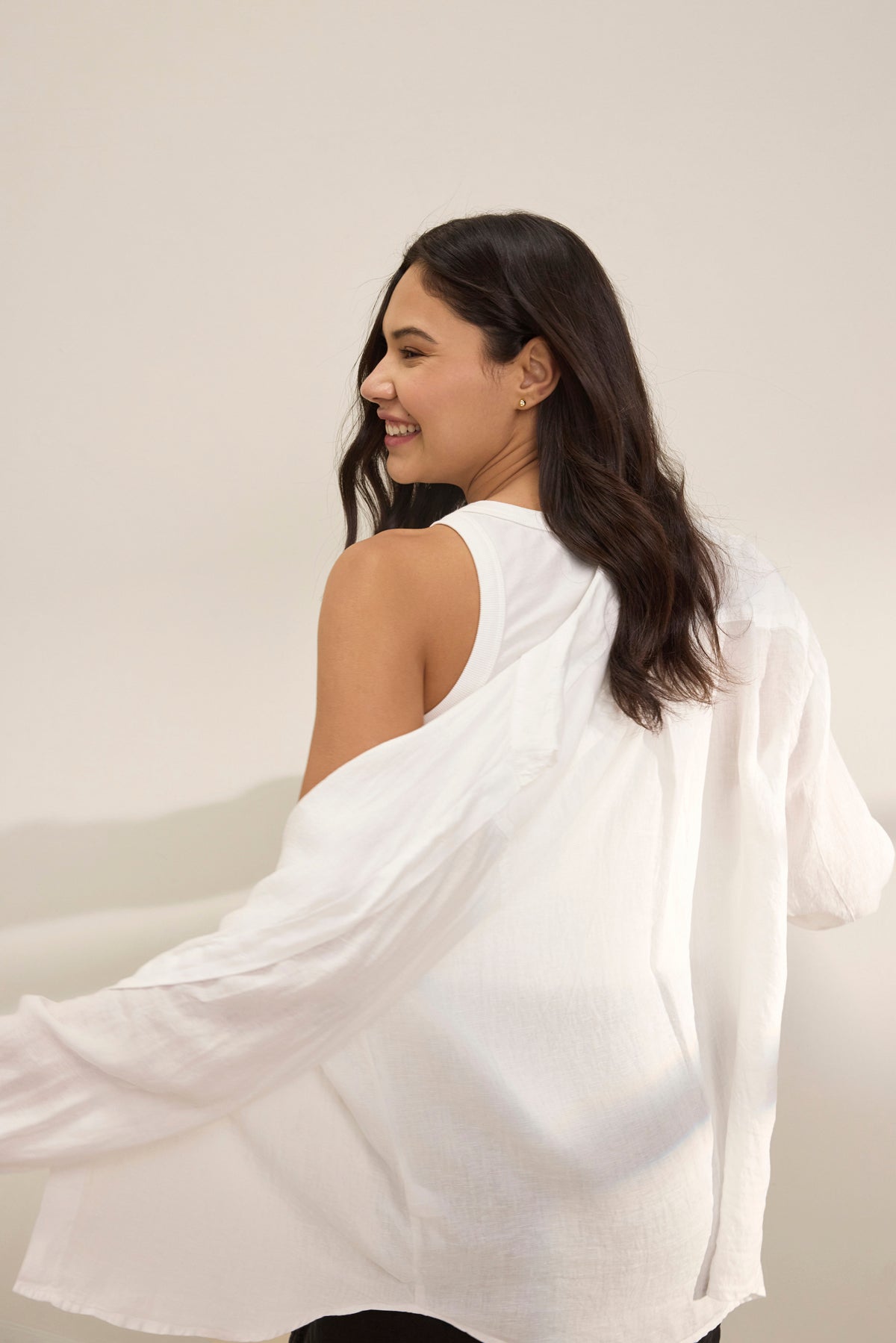   A woman with long dark hair, wearing a white sleeveless top and the SISI LINEN SHIRT by Velvet by Graham & Spencer, smiles while looking over her shoulder against a neutral background. 