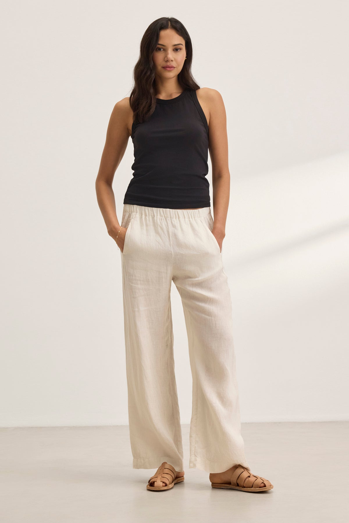   Against a neutral background, a person wears a black sleeveless top, Velvet by Graham & Spencer's STEPHANIE LINEN PANT in cream with a wide-leg design, and sandals. 