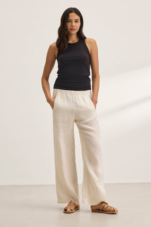 Against a neutral background, a person wears a black sleeveless top, Velvet by Graham & Spencer's STEPHANIE LINEN PANT in cream with a wide-leg design, and sandals.