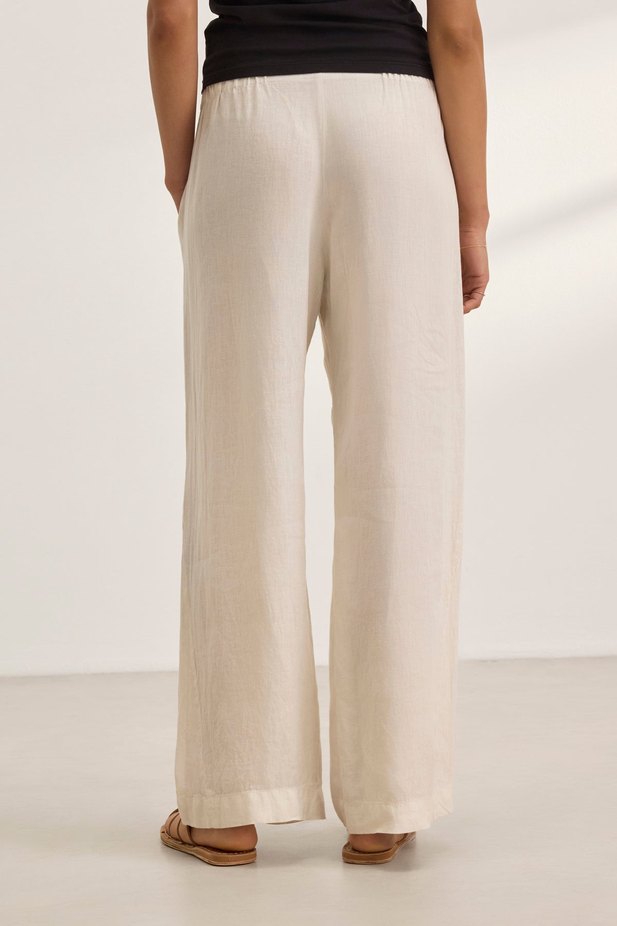   A person stands indoors against a neutral backdrop, wearing relaxed-fit STEPHANIE LINEN PANTS by Velvet by Graham & Spencer in beige with wide legs, paired with a sleek black top. 