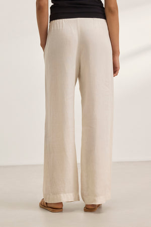 A person stands indoors against a neutral backdrop, wearing relaxed-fit STEPHANIE LINEN PANTS by Velvet by Graham & Spencer in beige with wide legs, paired with a sleek black top.