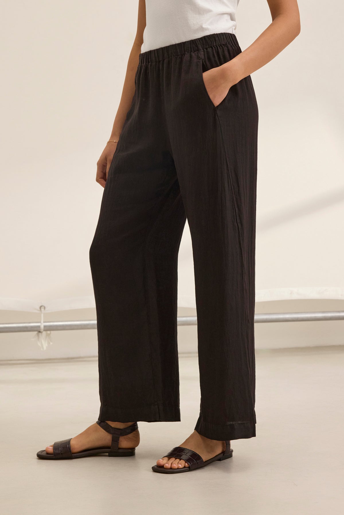   A person wearing a white top and Velvet by Graham & Spencer's STEPHANIE LINEN PANT, featuring a black loose fit with an elastic waist, stands indoors, comfortably sporting black sandals. 