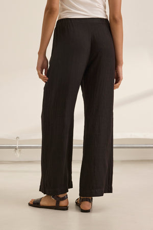 A person wears STEPHANIE LINEN PANT by Velvet by Graham & Spencer, showcasing a lightweight wide-leg design, paired with a white shirt and black sandals, viewed from behind.