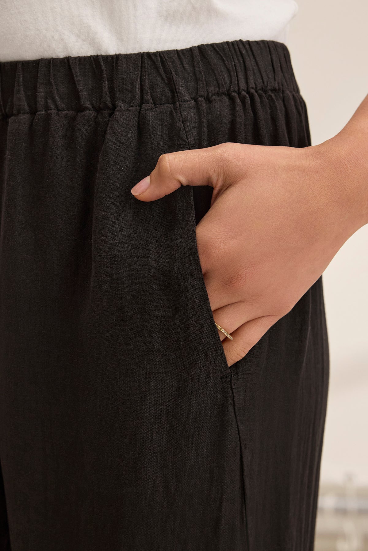   A person wears Velvet by Graham & Spencer's STEPHANIE LINEN PANT, featuring a black wide-leg and elastic waist, with their hand in the pocket. A gold ring adorns their finger. 