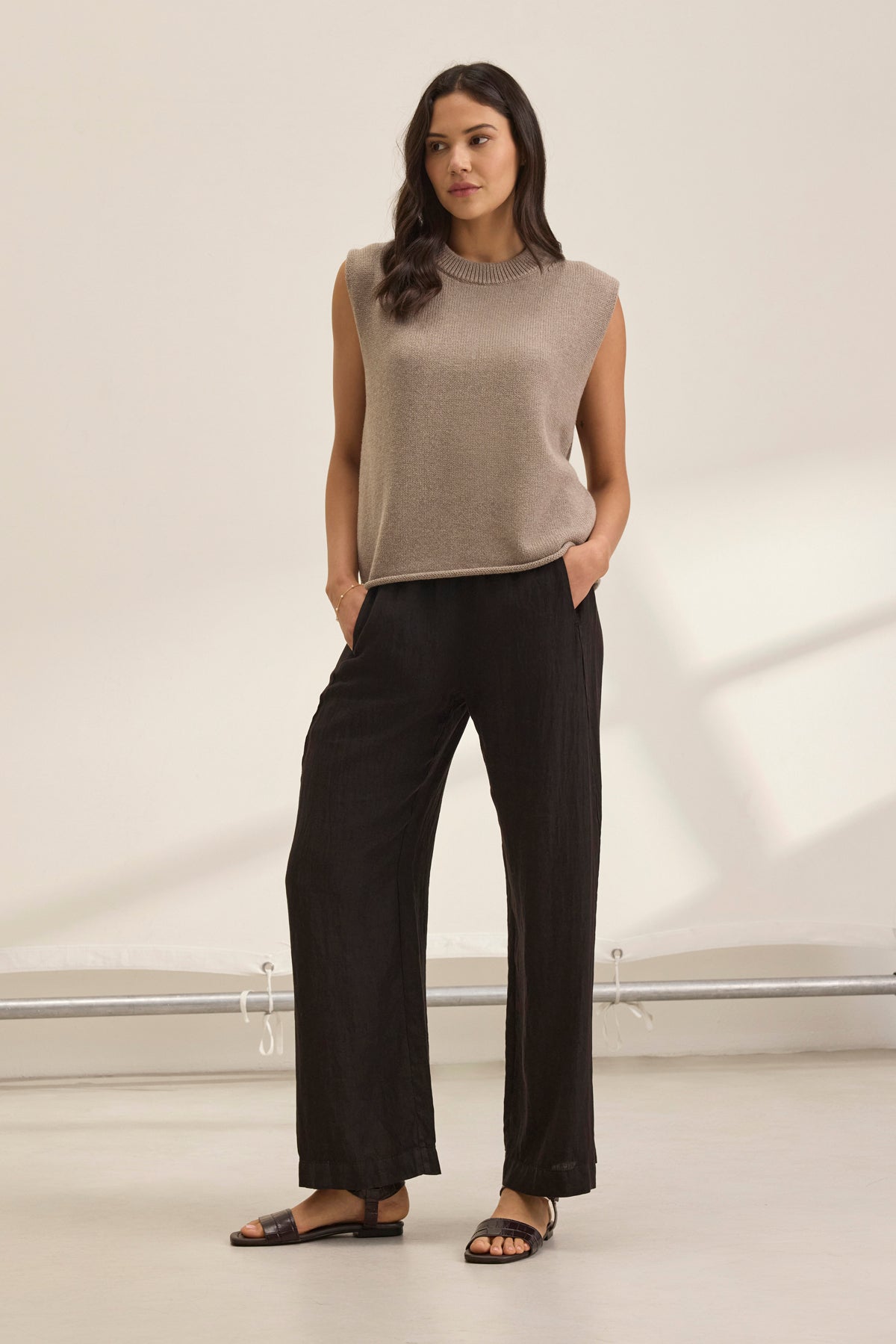   A person wearing the STEPHANIE LINEN PANT from Velvet by Graham & Spencer with a beige sleeveless top, stands with hands in pockets against a light background. 