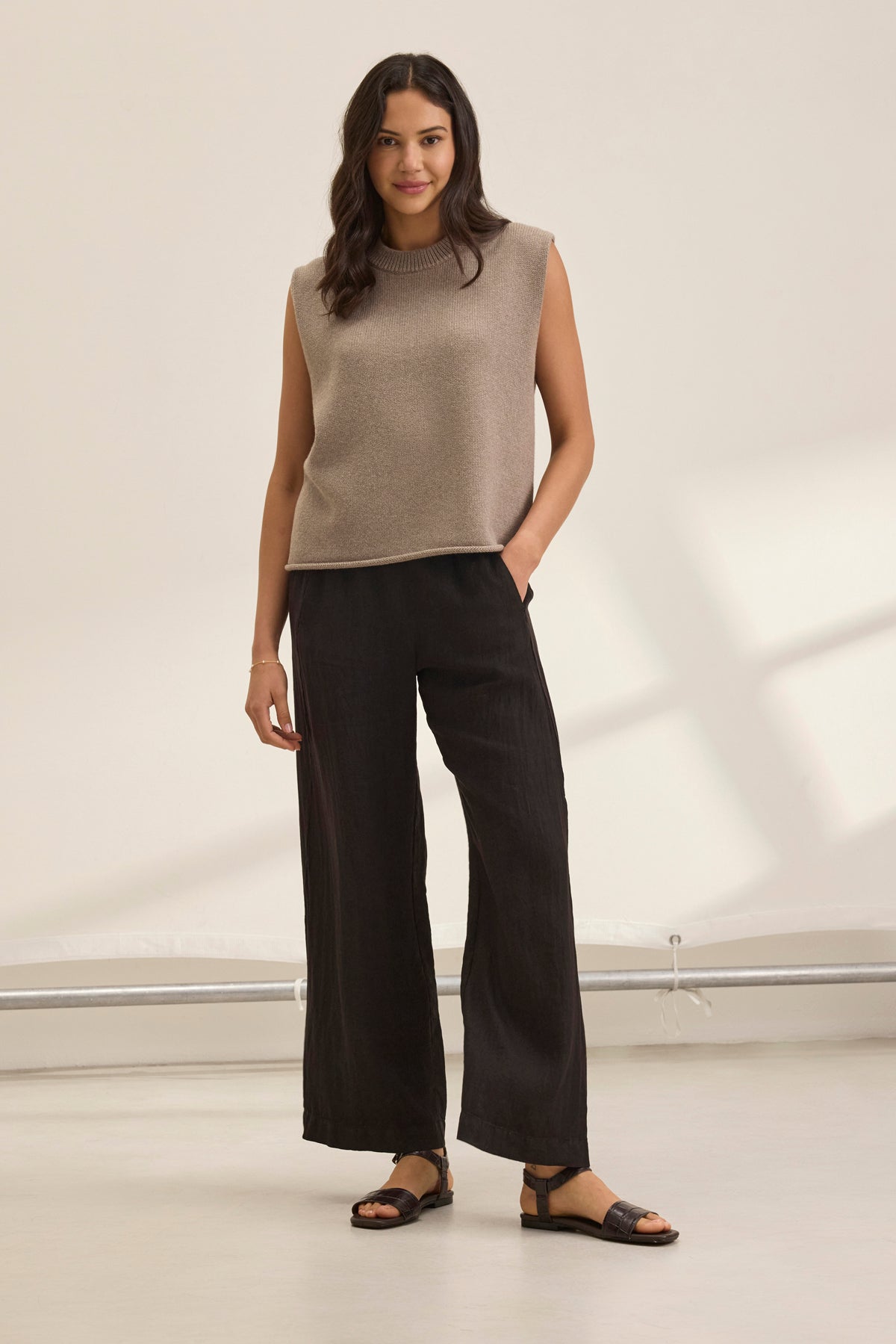   A woman smiles in a beige sleeveless top and the STEPHANIE LINEN PANT by Velvet by Graham & Spencer, featuring an elastic waist design, with sandals, posing against a plain white background. 