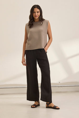 A woman smiles in a beige sleeveless top and the STEPHANIE LINEN PANT by Velvet by Graham & Spencer, featuring an elastic waist design, with sandals, posing against a plain white background.