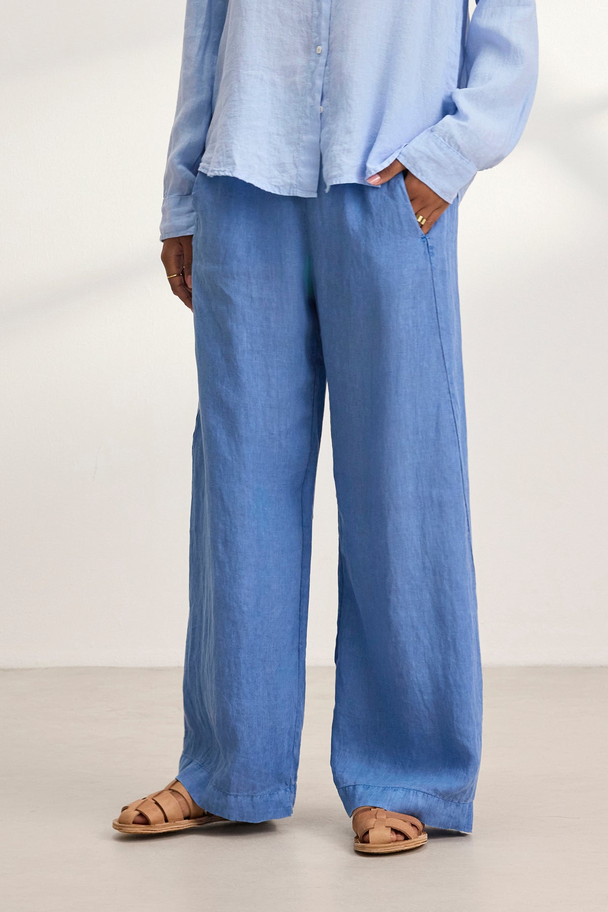 A person wearing a light blue shirt and the relaxed fit STEPHANIE LINEN PANT by Velvet by Graham & Spencer, with hands in pockets, stands on a light floor, sporting brown sandals.-38819778920641