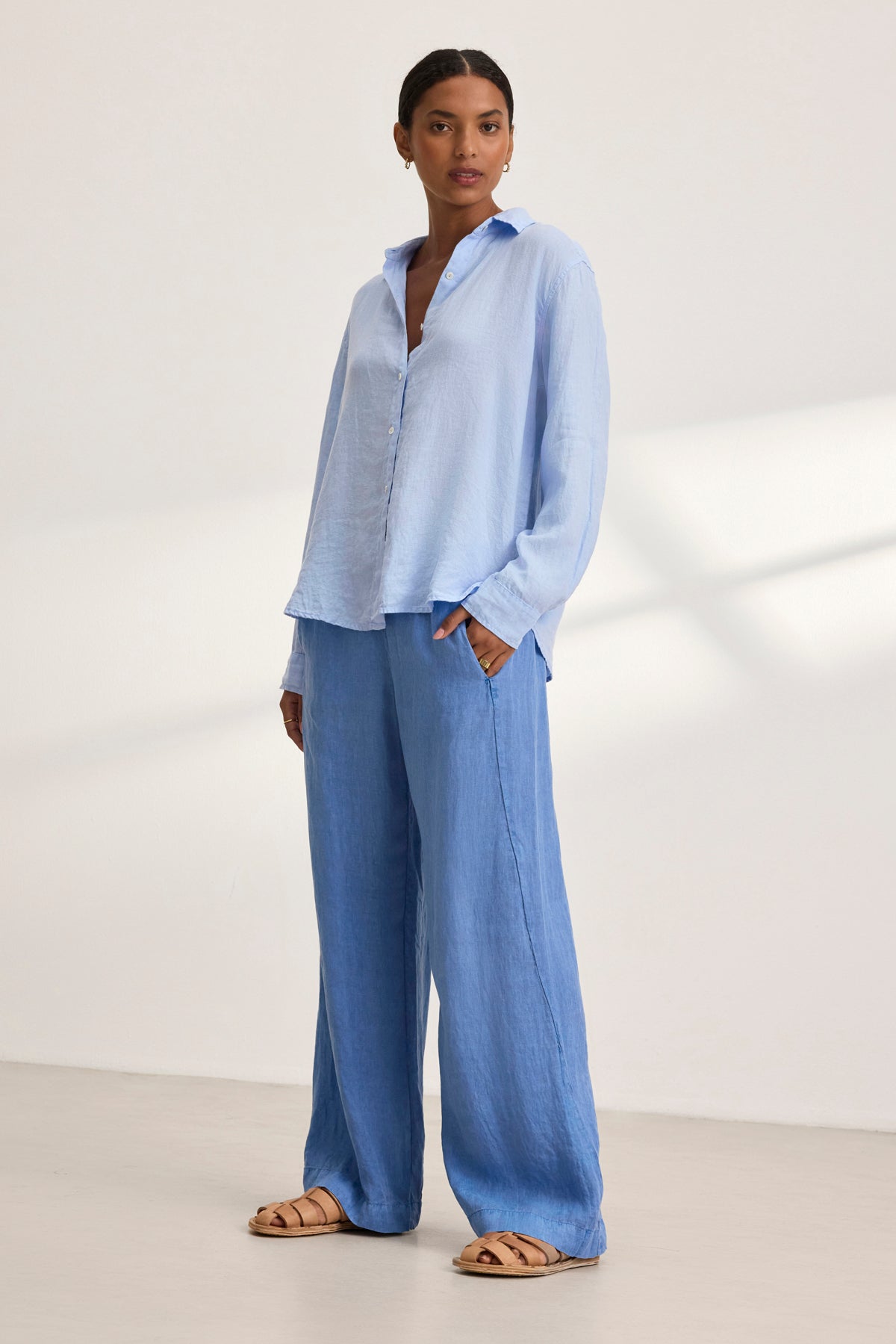   A person in a light blue shirt and Velvet by Graham & Spencer's STEPHANIE LINEN PANT, featuring a relaxed fit, stands against a plain backdrop. Hands are in pockets, complemented by sandals. 