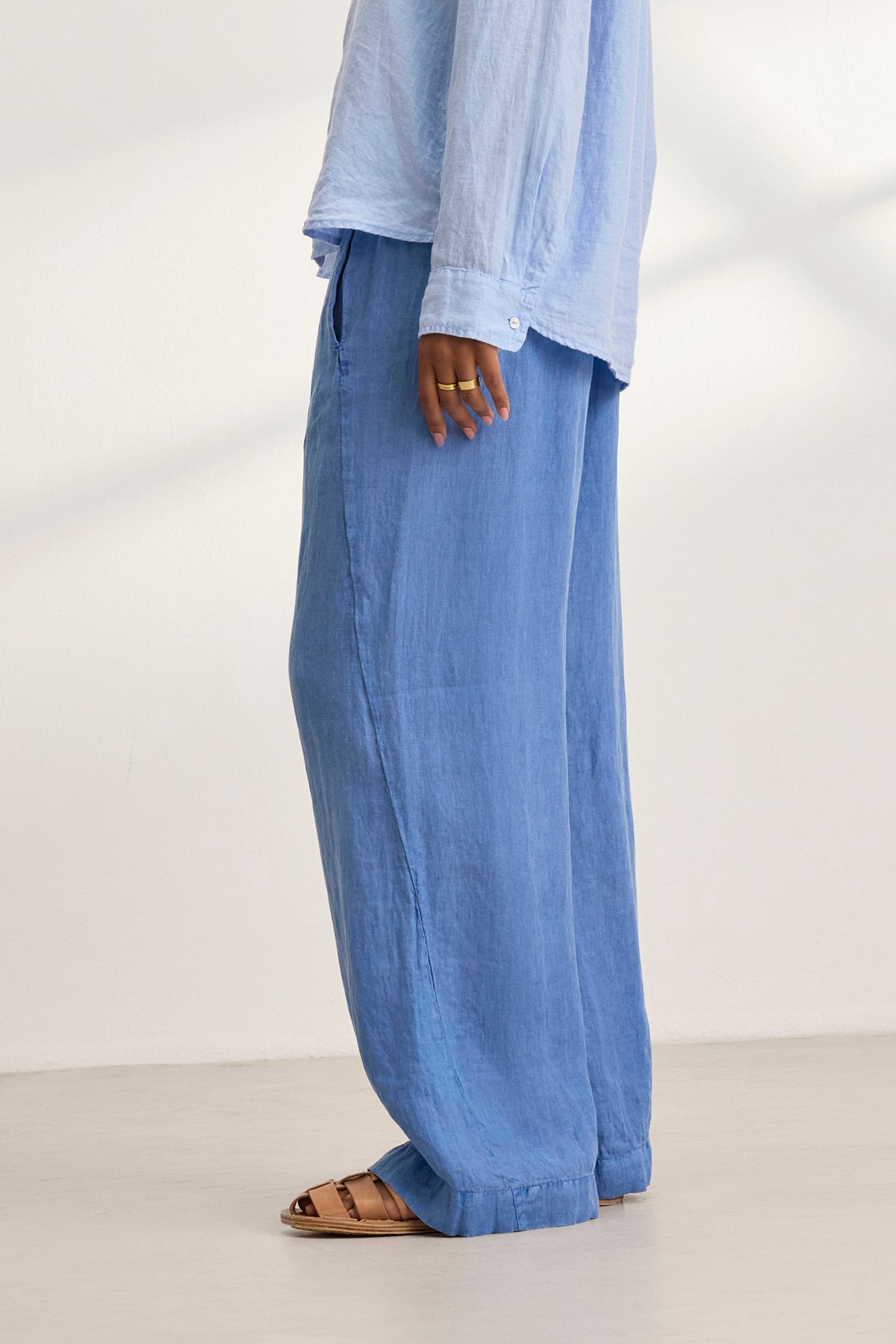   A person stands on a light floor, wearing the STEPHANIE LINEN PANT by Velvet by Graham & Spencer. The loose, light blue pants have a relaxed fit and wide-leg design, paired with a matching blouse. Brown sandals are visible on their feet. 