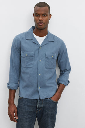 A man wearing the DONOVAN BUTTON-UP SHIRT in light blue cotton blend twill by Velvet by Graham & Spencer over a white T-shirt and jeans stands against a plain white background.