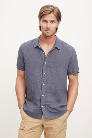 A man with shoulder-length hair stands with his hands in his pockets, wearing a short-sleeve MACKIE LINEN BUTTON-UP SHIRT by Velvet by Graham & Spencer and tan pants, perfect for a summer wardrobe.