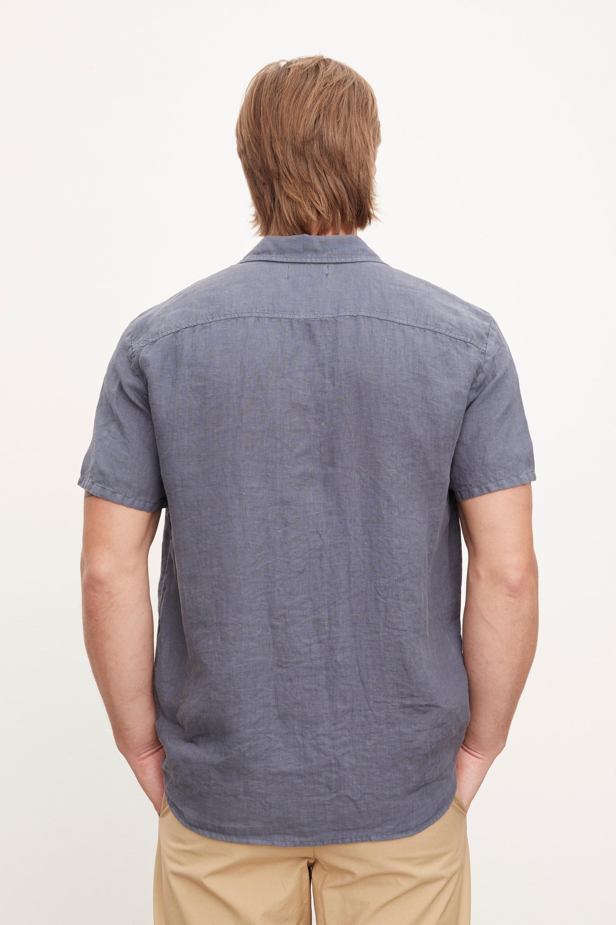   A person with shoulder-length blond hair is shown from the back, wearing a MACKIE LINEN BUTTON-UP SHIRT by Velvet by Graham & Spencer and beige pants, perfect for a summer wardrobe. 