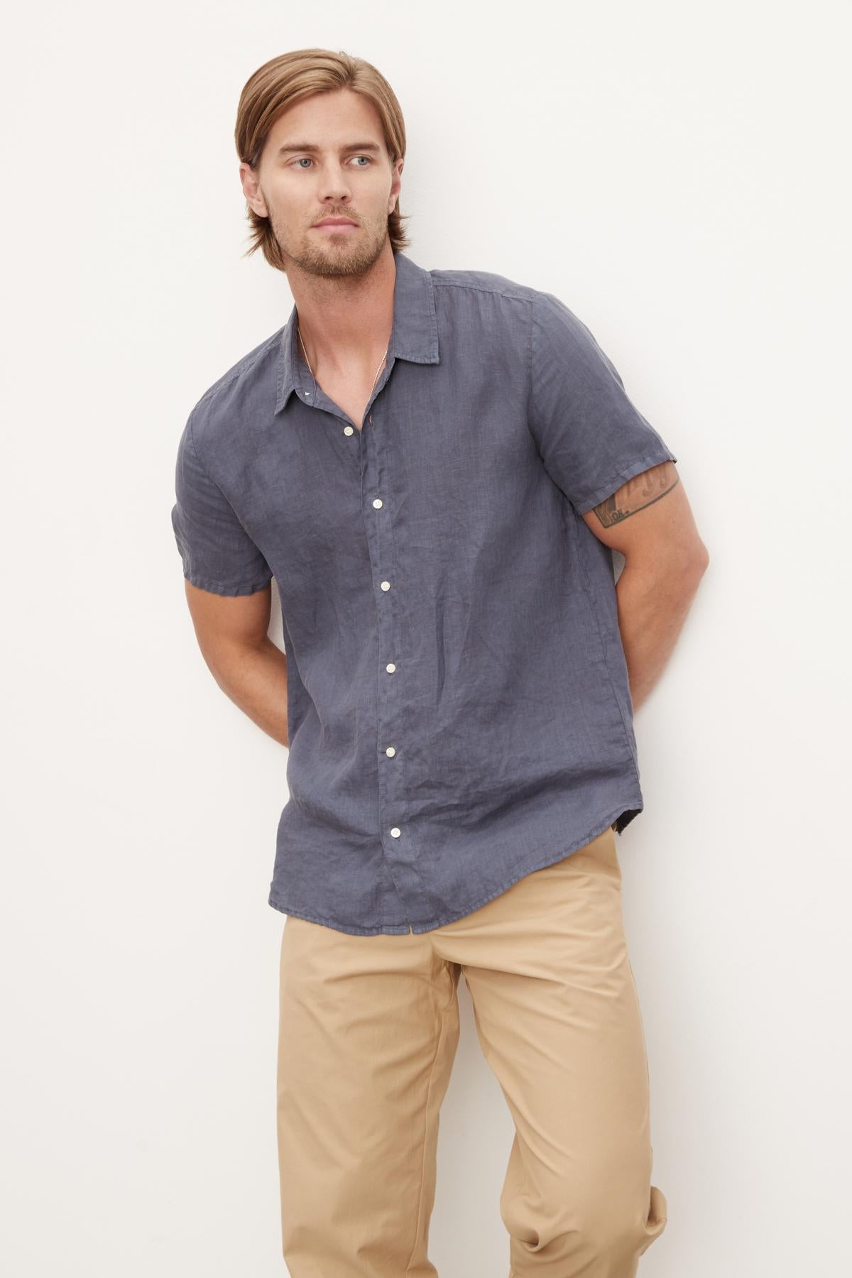   A man wearing a blue MACKIE LINEN BUTTON-UP SHIRT from Velvet by Graham & Spencer and beige pants stands against a plain white background, embodying the perfect summer wardrobe. 