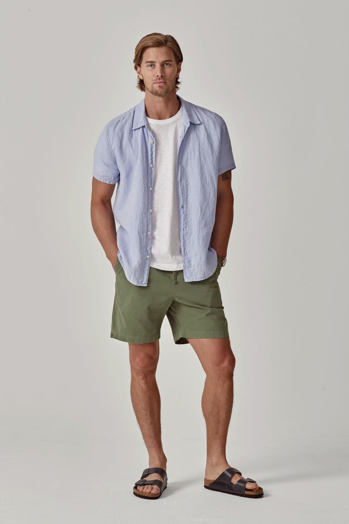   A man with shoulder-length hair stands wearing a Velvet by Graham & Spencer MACKIE LINEN BUTTON-UP SHIRT over a white t-shirt, green shorts, and black sandals, hands in pockets. 